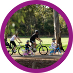 A round photo of a family riding bikes through the park past gumtrees and sunlit lawn